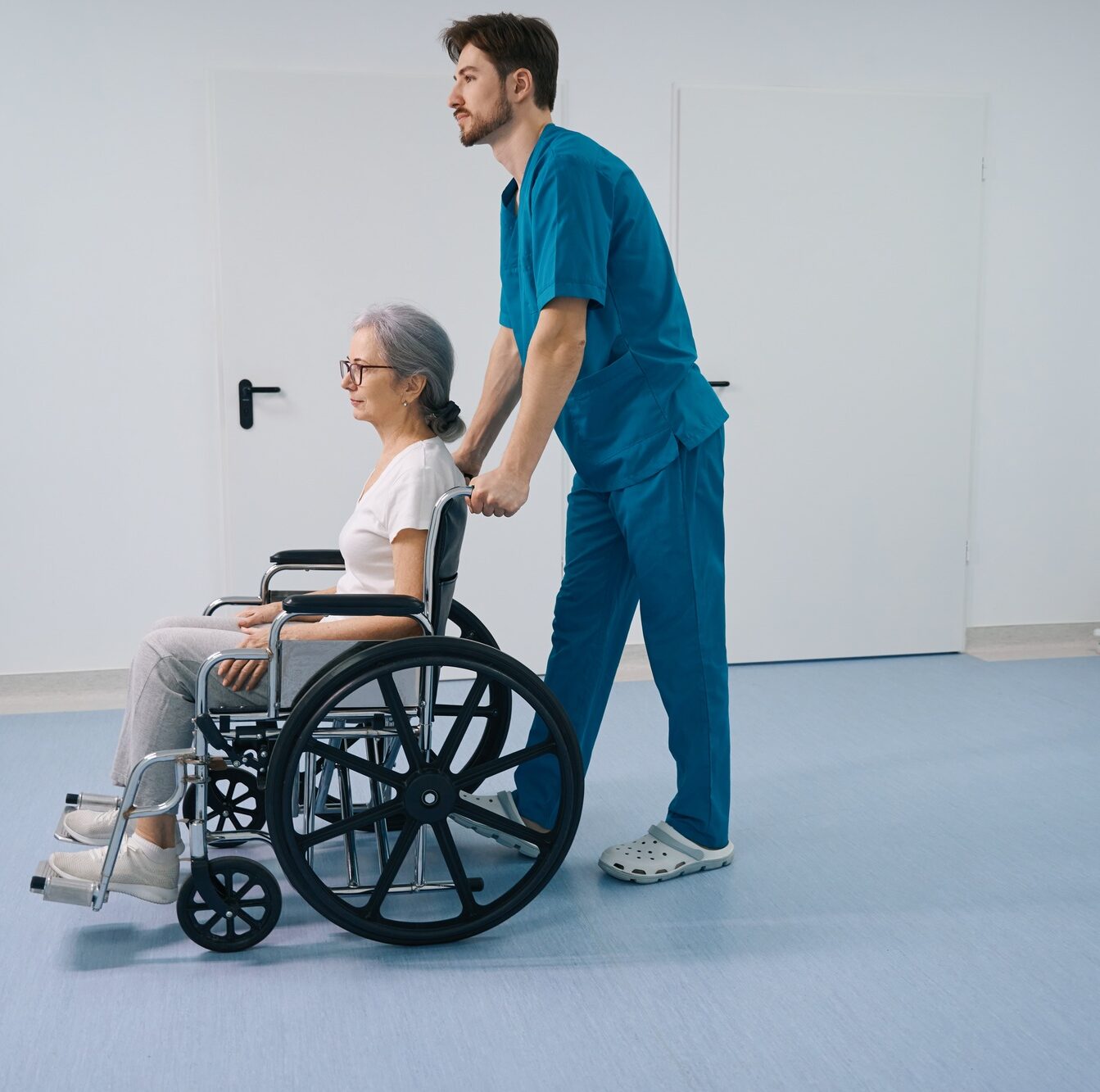 Caring nurse is carrying an elderly lady in a wheelchair in hospital corridor